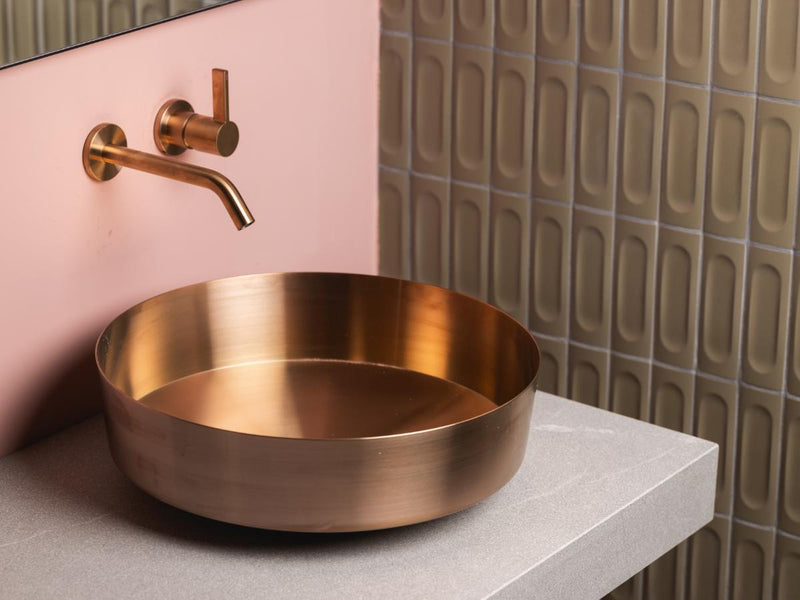 A round brushed copper vessel basin from LAUFEN mounted on a light gray stone countertop, paired with a matching wall-mounted copper faucet against a pink wall and textured beige tiles.