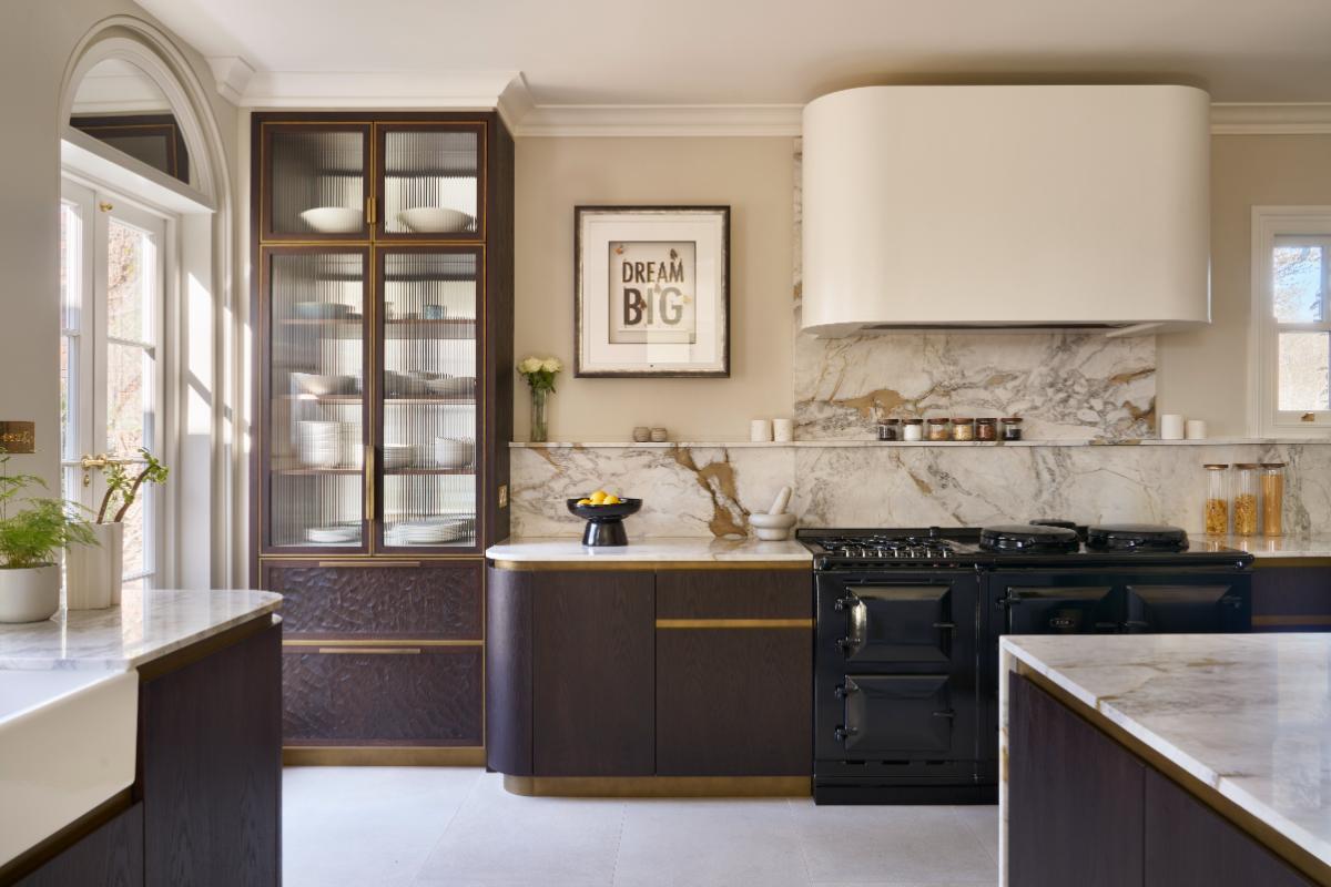 A luxury kitchen by Ledbury Studio featuring dark wood cabinetry with gold handles, a black freestanding range, a white marble backsplash, a large white range hood, and a central framed print that reads 'DREAM BIG'.