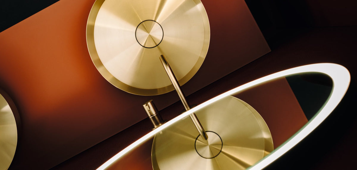 LAUFEN Rivo washbasin and wall spout in brushed brass, seen from above against terracotta, with a lit ring mirror